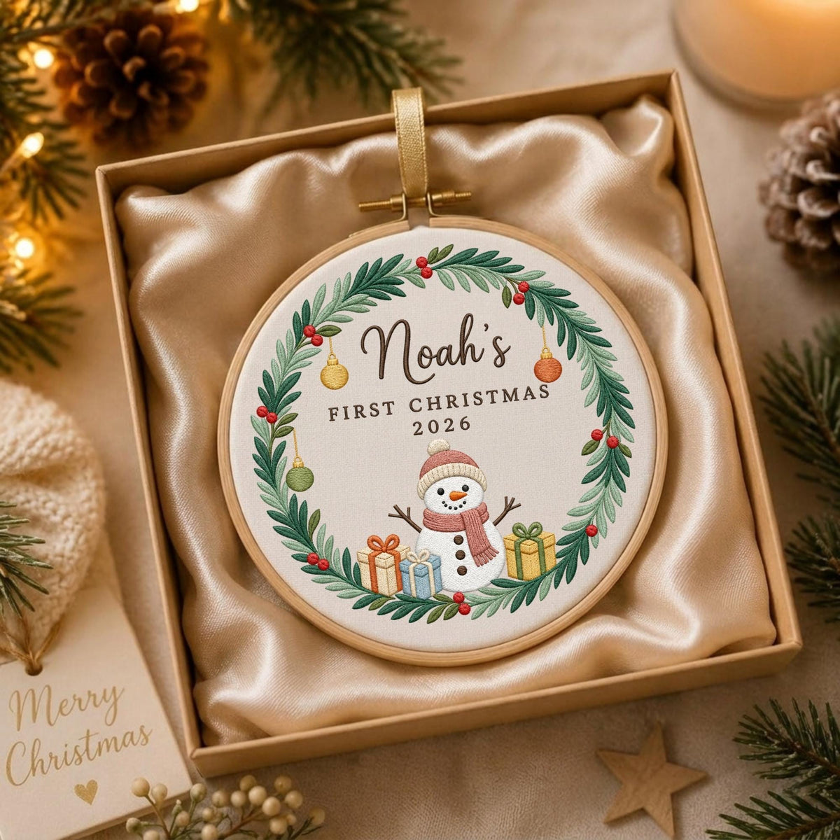 May include: A round embroidery hoop ornament featuring a white fabric center with a green wreath, a snowman wearing a pink scarf, and colorful gift boxes. The text reads Noah's FIRST CHRISTMAS 2026. The ornament is displayed in a gift box.