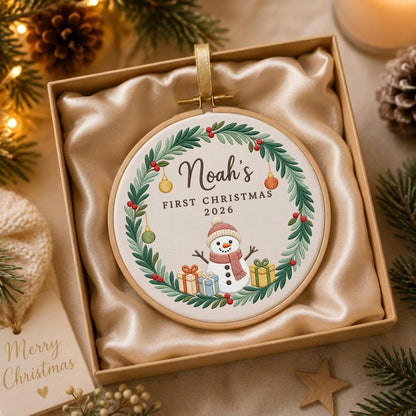 May include: A round embroidery hoop ornament featuring a white fabric center with a green wreath, a snowman wearing a pink scarf, and colorful gift boxes. The text reads Noah's FIRST CHRISTMAS 2026. The ornament is displayed in a gift box.