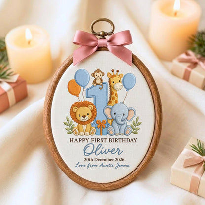 May include: An oval wooden embroidery hoop frame containing a fabric panel with a printed illustration of a lion, giraffe, elephant, and monkey around a large blue number one. Text reads: HAPPY FIRST BIRTHDAY, Oliver, 20th December 2026, Love from Auntie Jemma. A pink satin bow is attached to the top.