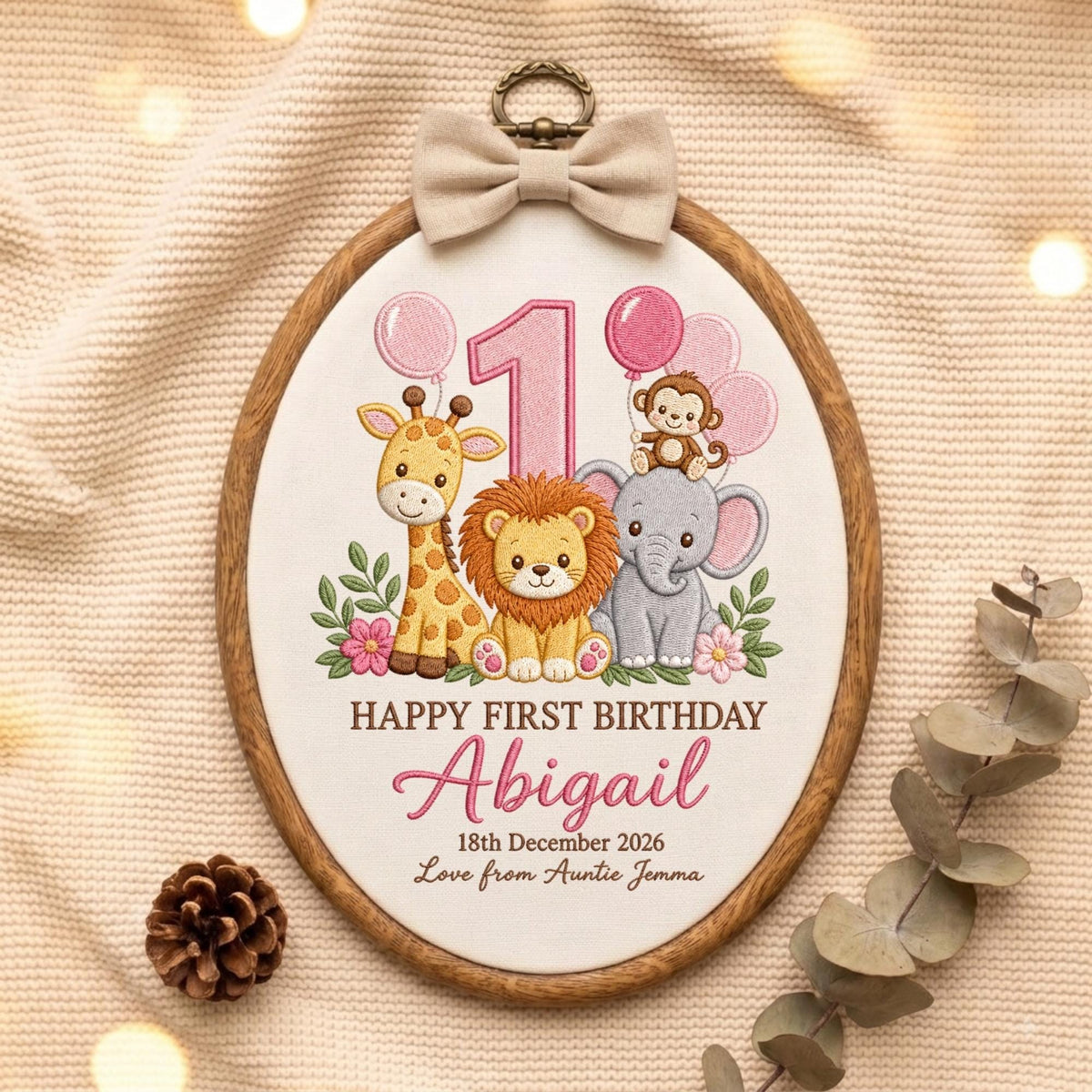 May include: An oval embroidery hoop featuring a white fabric center with a giraffe, lion, elephant, and monkey illustration. Text reads: HAPPY FIRST BIRTHDAY Abigail 18th December 2026 Love from Auntie Jemma. A beige bow is attached to the top.