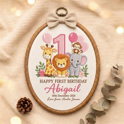 May include: An oval embroidery hoop featuring a white fabric center with a giraffe, lion, elephant, and monkey illustration. Text reads: HAPPY FIRST BIRTHDAY Abigail 18th December 2026 Love from Auntie Jemma. A beige bow is attached to the top.