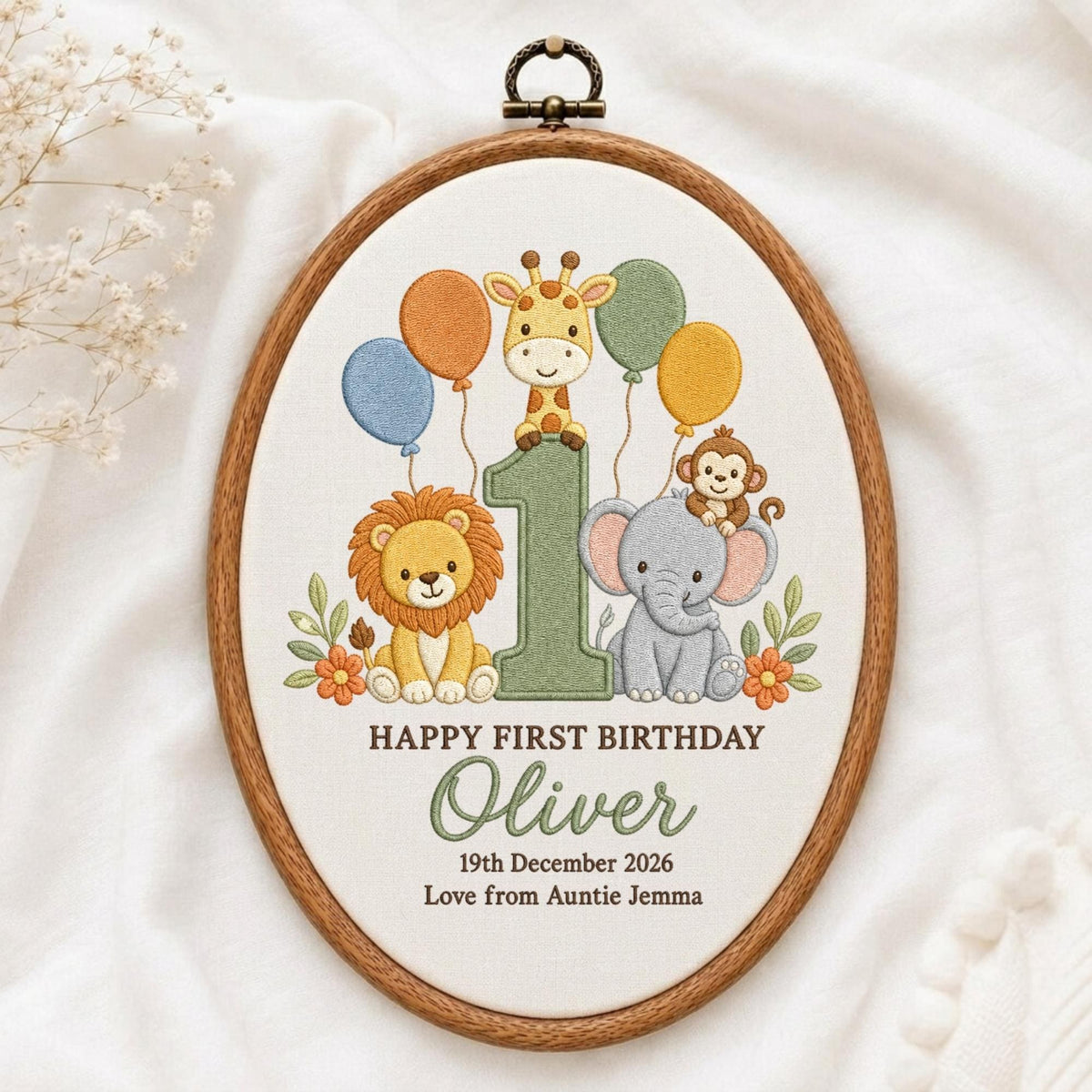 May include: An oval embroidery hoop containing fabric with a stitched design of a giraffe, lion, monkey, and elephant around a large number one. Text reads: HAPPY FIRST BIRTHDAY, Oliver, 19th December 2026, Love from Auntie Jemma.