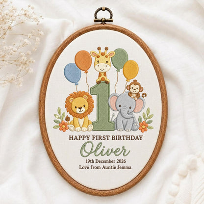 May include: An oval embroidery hoop containing fabric with a stitched design of a giraffe, lion, monkey, and elephant around a large number one. Text reads: HAPPY FIRST BIRTHDAY, Oliver, 19th December 2026, Love from Auntie Jemma.