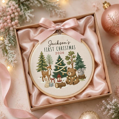 May include: A round wooden embroidery hoop featuring a cream-colored fabric with a forest scene, including a deer, bear, rabbit, and squirrel. The text reads Jackson's FIRST CHRISTMAS 2026. A light pink satin ribbon bow is attached to the top of the hoop.