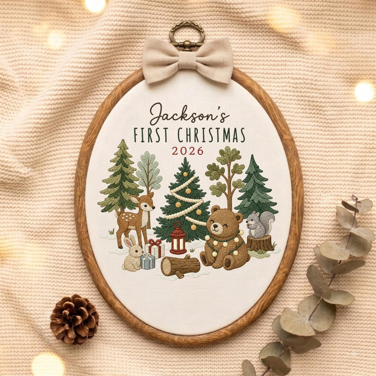 May include: An oval embroidery hoop featuring a woodland scene with a deer, rabbit, bear, and squirrel among pine trees, gifts, and a lantern. The text reads Jackson's FIRST CHRISTMAS 2026. A beige fabric bow is attached to the top metal hanger.