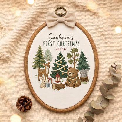 May include: An oval embroidery hoop featuring a woodland scene with a deer, rabbit, bear, and squirrel among pine trees, gifts, and a lantern. The text reads Jackson's FIRST CHRISTMAS 2026. A beige fabric bow is attached to the top metal hanger.