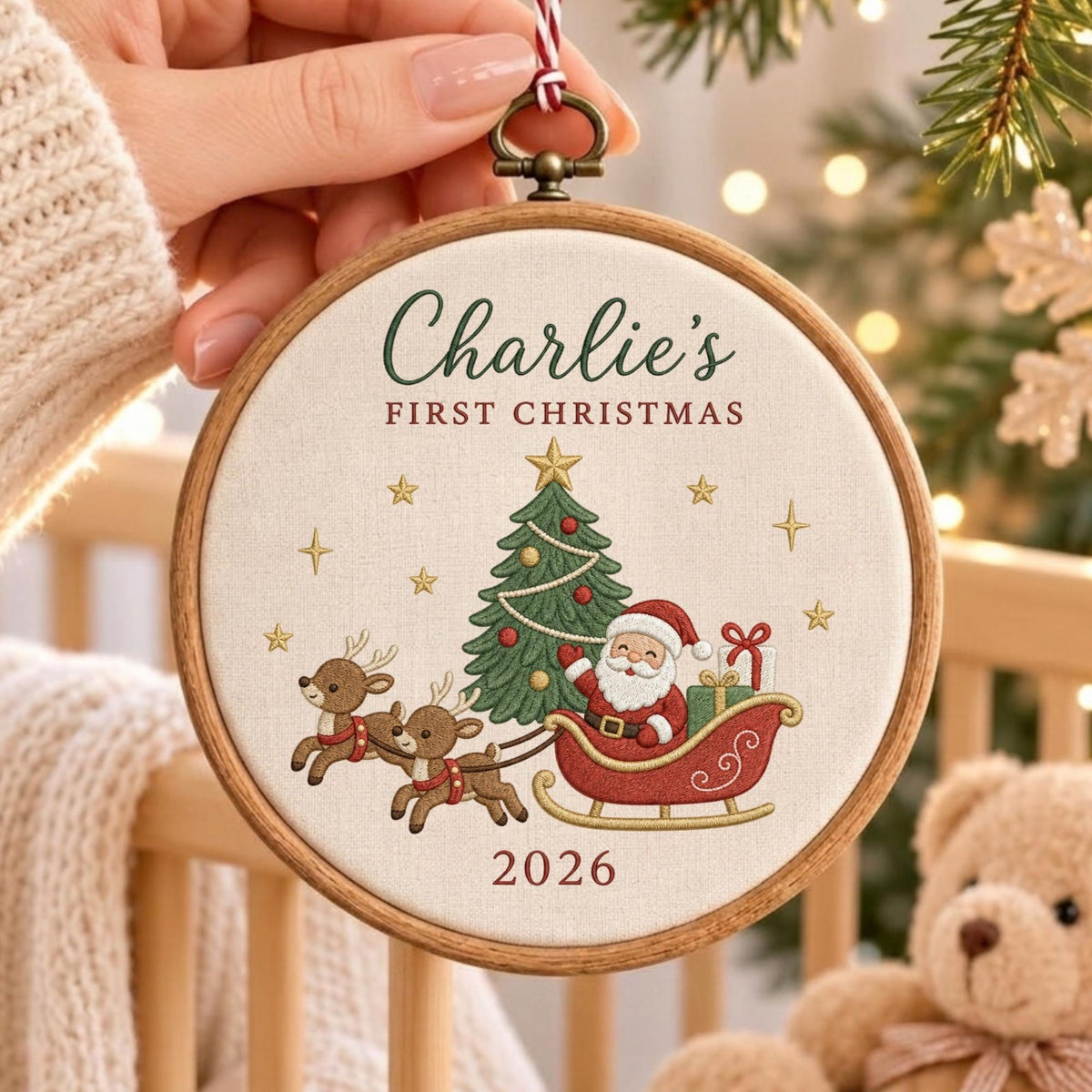 May include: A round wooden embroidery hoop ornament featuring off-white fabric with embroidered text Charlie's FIRST CHRISTMAS and 2026. The design includes a green Christmas tree, a red sleigh with Santa Claus, and two brown reindeer.