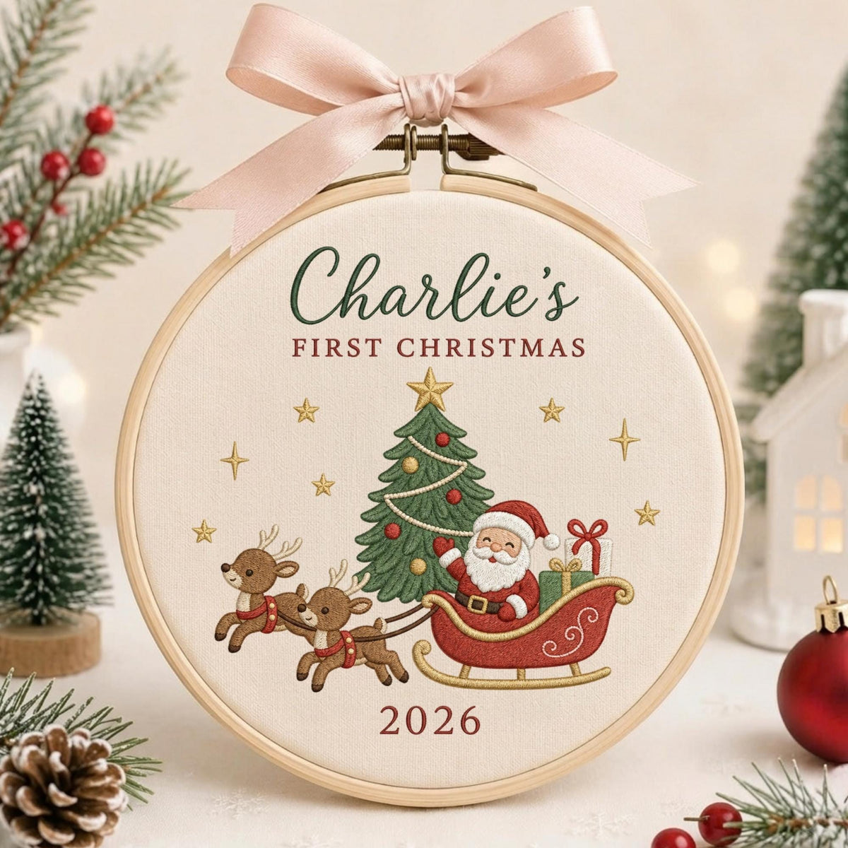 May include: A round wooden embroidery hoop containing cream-colored fabric with embroidered text reading Charlie's FIRST CHRISTMAS and 2026. The design features a green Christmas tree, two brown reindeer, and Santa Claus in a red sleigh, topped with a pink ribbon.
