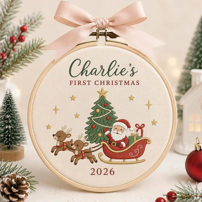 May include: A round wooden embroidery hoop containing cream-colored fabric with embroidered text reading Charlie's FIRST CHRISTMAS and 2026. The design features a green Christmas tree, two brown reindeer, and Santa Claus in a red sleigh, topped with a pink ribbon.