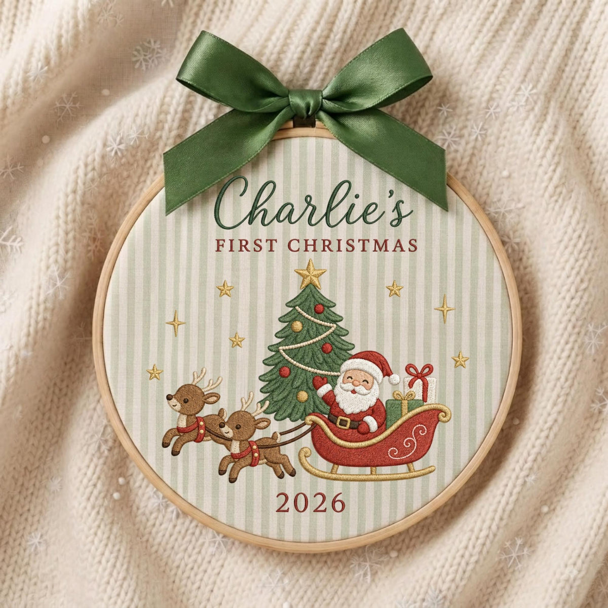 May include: A round wooden embroidery hoop ornament featuring a light green and white striped fabric background. The text reads Charlie's FIRST CHRISTMAS 2026. It depicts a Christmas tree, Santa in a red sleigh, and two reindeer, topped with a dark green satin bow.