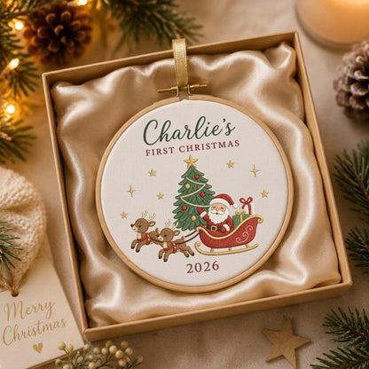 May include: A round wooden embroidery hoop ornament featuring a cream fabric center with an embroidered design of Santa Claus in a sleigh pulled by two reindeer, a Christmas tree, and the text Charlie's FIRST CHRISTMAS 2026 in a gift box.