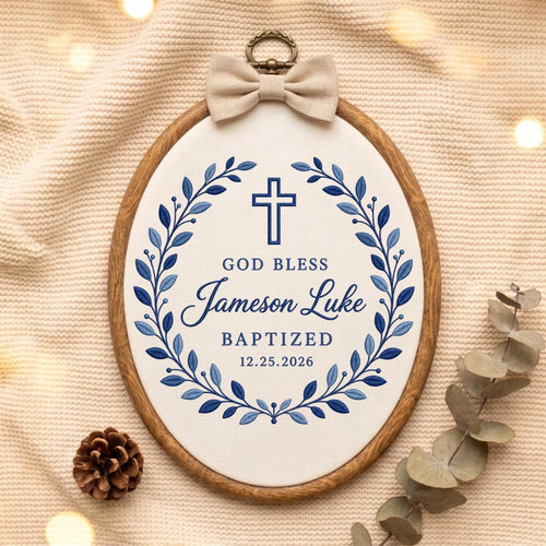 May include: An oval wooden embroidery hoop containing white fabric with blue embroidered text reading GOD BLESS Jameson Luke BAPTIZED 12.25.2026, a blue cross, and a blue leaf wreath. A beige fabric bow is attached to the top metal hanging loop.