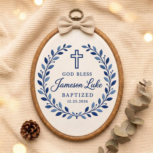 May include: An oval wooden embroidery hoop containing white fabric with blue embroidered text reading GOD BLESS Jameson Luke BAPTIZED 12.25.2026, a blue cross, and a blue leaf wreath. A beige fabric bow is attached to the top metal hanging loop.