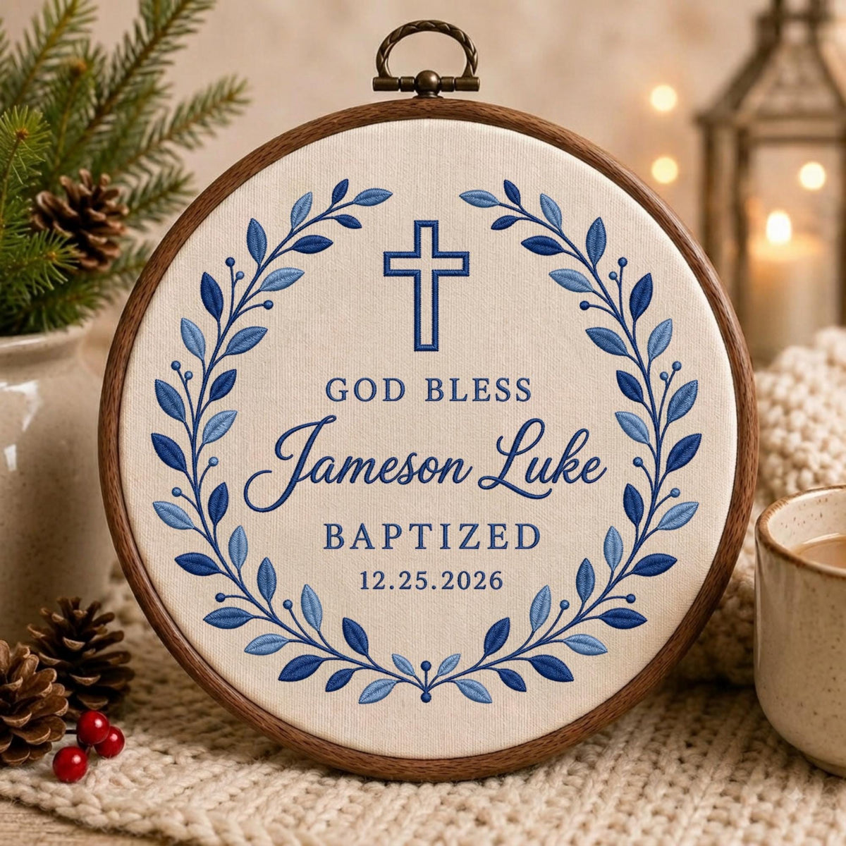May include: A circular wooden embroidery hoop containing cream-colored fabric with blue embroidered text reading GOD BLESS Jameson Luke BAPTIZED 12.25.2026. A blue cross sits above the text, framed by a symmetrical blue leaf wreath design.