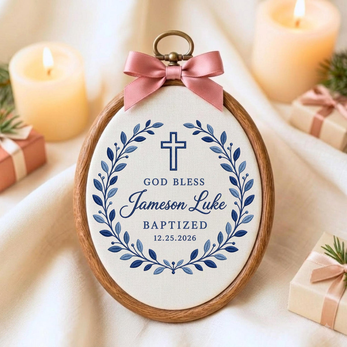 May include: An oval wooden embroidery hoop frame containing white fabric with a blue printed design of a cross, a leaf wreath, and the text GOD BLESS Jameson Luke BAPTIZED 12.25.2026. A dusty pink satin ribbon bow is attached to the top metal hanger.