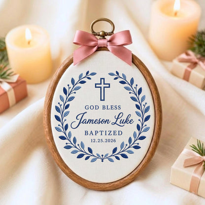 May include: An oval wooden embroidery hoop frame containing white fabric with a blue printed design of a cross, a leaf wreath, and the text GOD BLESS Jameson Luke BAPTIZED 12.25.2026. A dusty pink satin ribbon bow is attached to the top metal hanger.