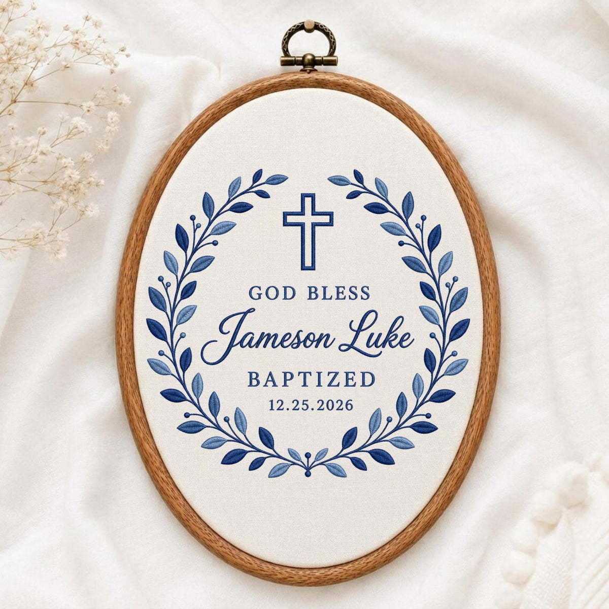 May include: An oval embroidery hoop frame containing white fabric with a blue embroidered wreath design. Centered inside is a blue cross and the text: GOD BLESS, Jameson Luke, BAPTIZED, 12.25.2026. The wooden frame features a metal hanging loop at the top.