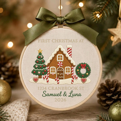 May include: A round embroidery hoop ornament featuring a gingerbread house, a Christmas tree, and a wreath on cream fabric. Text reads: FIRST CHRISTMAS AT, 1234 CRANBROOK ST, Samuel &amp; Luna, 2026. An olive green satin ribbon is tied at the top.