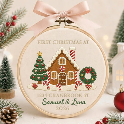 May include: A round wooden embroidery hoop featuring a cream fabric background with an embroidered gingerbread house, a Christmas tree, and a wreath. The text reads FIRST CHRISTMAS AT 1234 CRANBROOK ST SAMUEL &amp; LUNA 2026. A light pink ribbon is tied at the top.