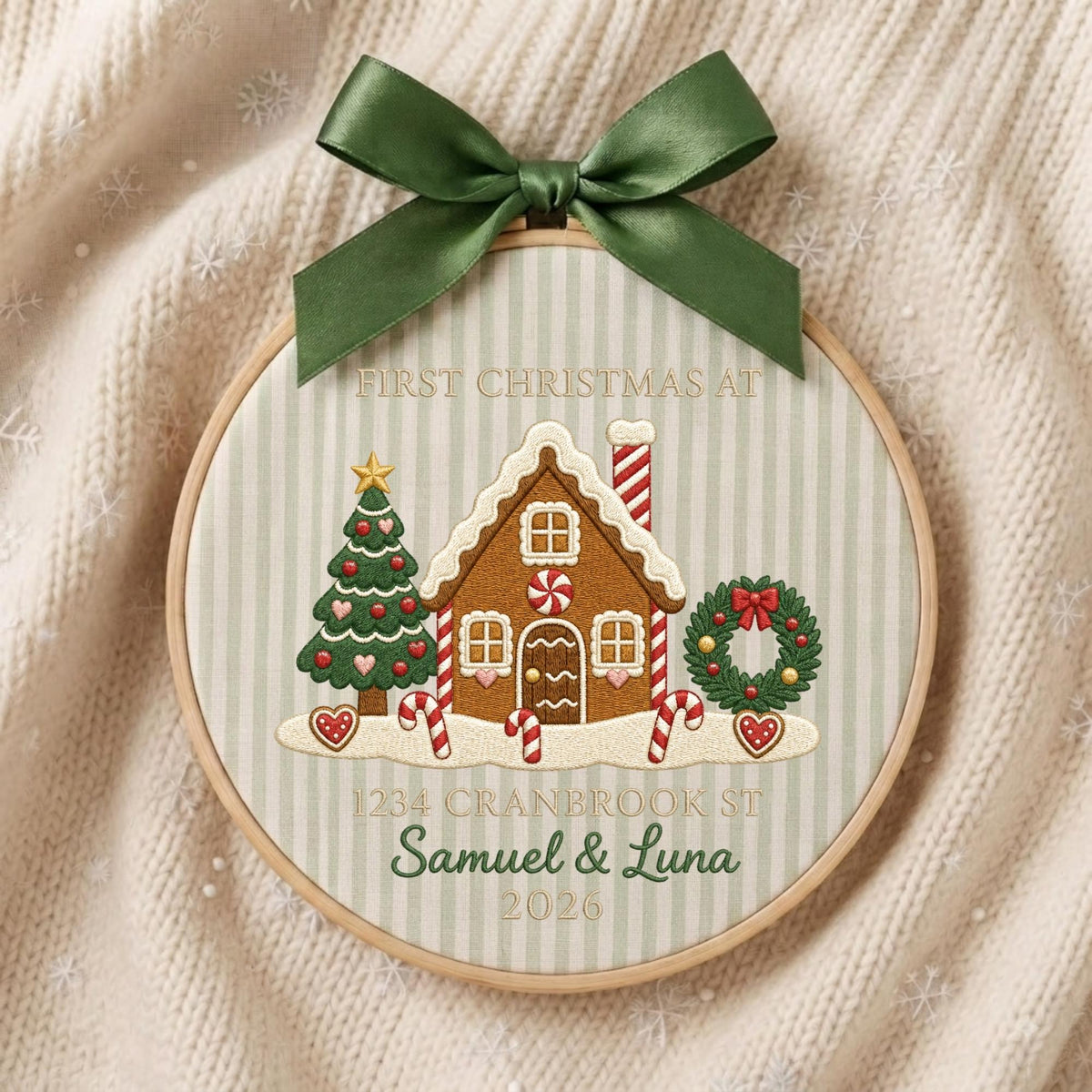 May include: A circular embroidery hoop ornament featuring a gingerbread house, a Christmas tree, and a wreath. The fabric has light green stripes and text reading FIRST CHRISTMAS AT 1234 CRANBROOK ST Samuel &amp; Luna 2026. A dark green satin bow is attached.