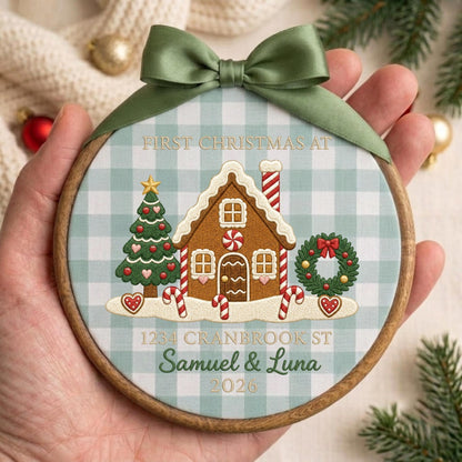 May include: A round wooden embroidery hoop ornament featuring a light blue and white gingham fabric background. It displays a gingerbread house, a Christmas tree, and a wreath with the text FIRST CHRISTMAS AT 1234 CRANBROOK ST SAMUEL &amp; LUNA 2026.