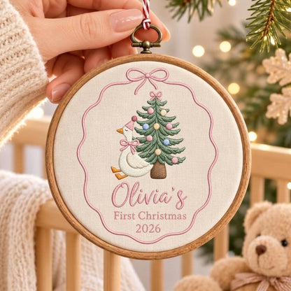 May include: A round wooden embroidery hoop ornament featuring a white fabric center with a pink embroidered border and bow. The design includes a white goose wearing a pink party hat, a decorated Christmas tree, and the text Olivia's First Christmas 2026.