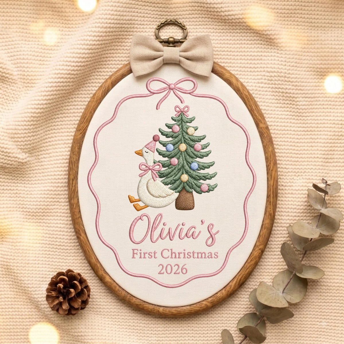 May include: An oval wooden embroidery hoop containing white fabric with a stitched design of a white goose wearing a pink hat and bow next to a decorated Christmas tree. The text reads Olivia's First Christmas 2026. A beige bow is attached.