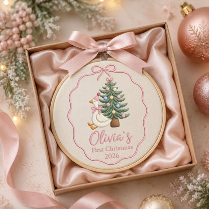 May include: A round wooden embroidery hoop containing cream fabric with a stitched design of a goose wearing a pink hat next to a Christmas tree. The text reads Olivia's First Christmas 2026. A pink satin ribbon is tied at the top of the hoop.