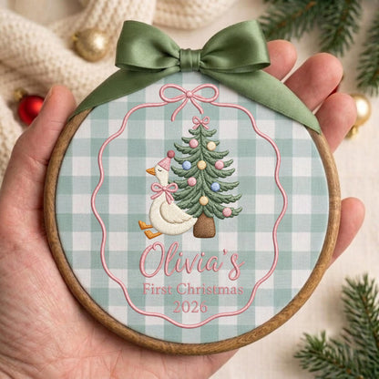 May include: A round wooden embroidery hoop ornament featuring a light green and white gingham fabric background. The center displays an embroidered white goose wearing a pink party hat and bow, next to a decorated Christmas tree. Text reads Olivia's First Christmas 2026. A sage green satin ribbon bow is tied at the top.
