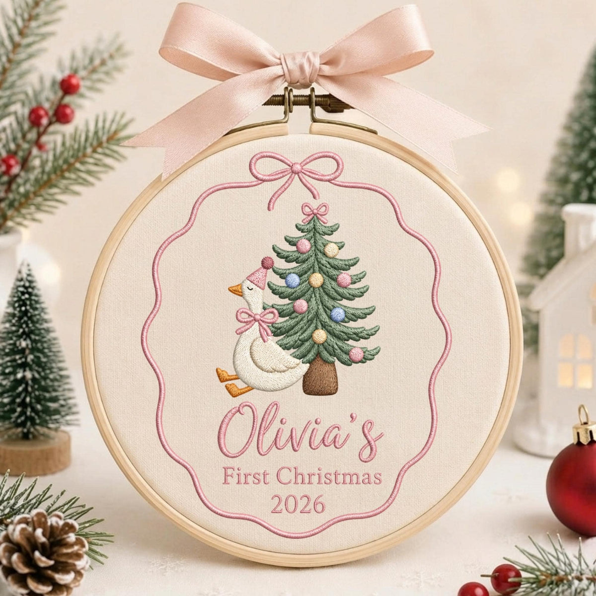 May include: A circular wooden embroidery hoop containing cream fabric with an embroidered white goose wearing a pink hat and bow next to a decorated Christmas tree. The text reads Olivia's First Christmas 2026. A pink ribbon bow is tied at the top.