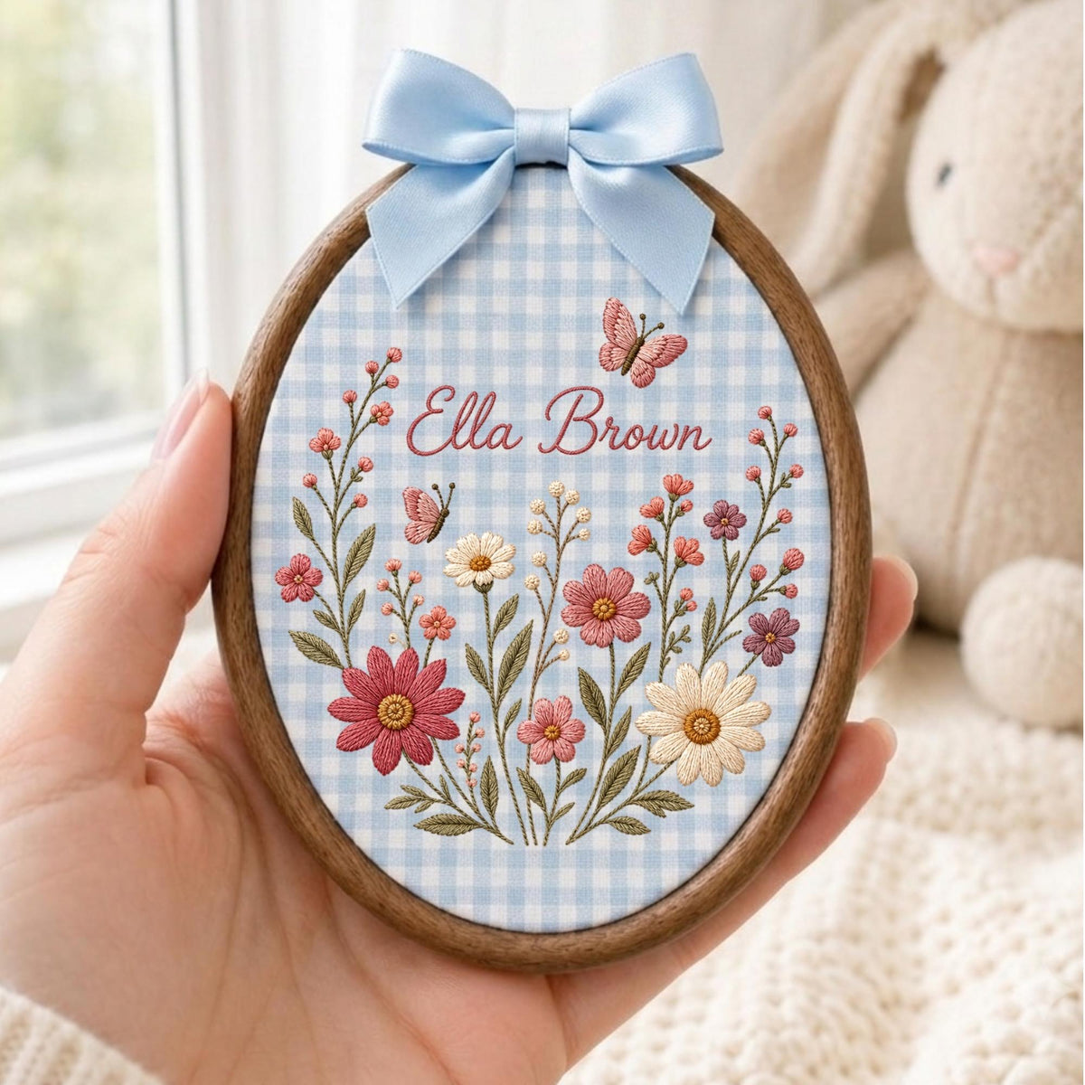 May include: An oval wooden embroidery hoop featuring a light blue and white gingham fabric background. The fabric is embroidered with colorful wildflowers, two small butterflies, and the name Ella Brown in a script font, topped with a light blue bow.