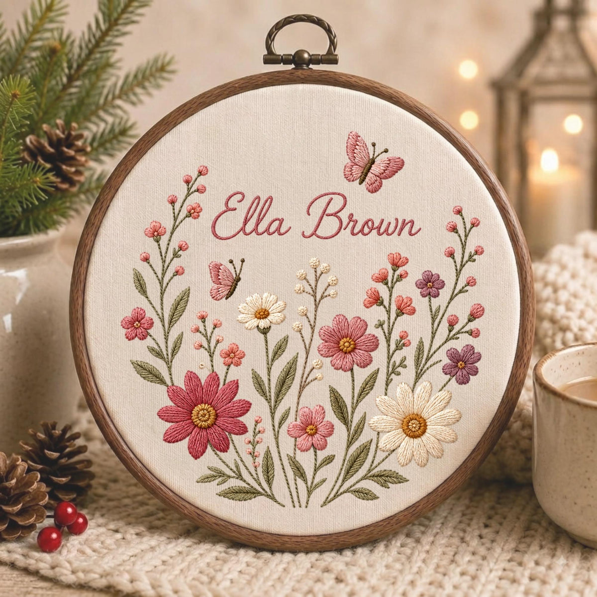 May include: A circular wooden embroidery hoop containing off-white fabric with a hand-stitched floral design. The embroidery features pink and white wildflowers, green stems, and two small pink butterflies. The name Ella Brown is stitched in pink.