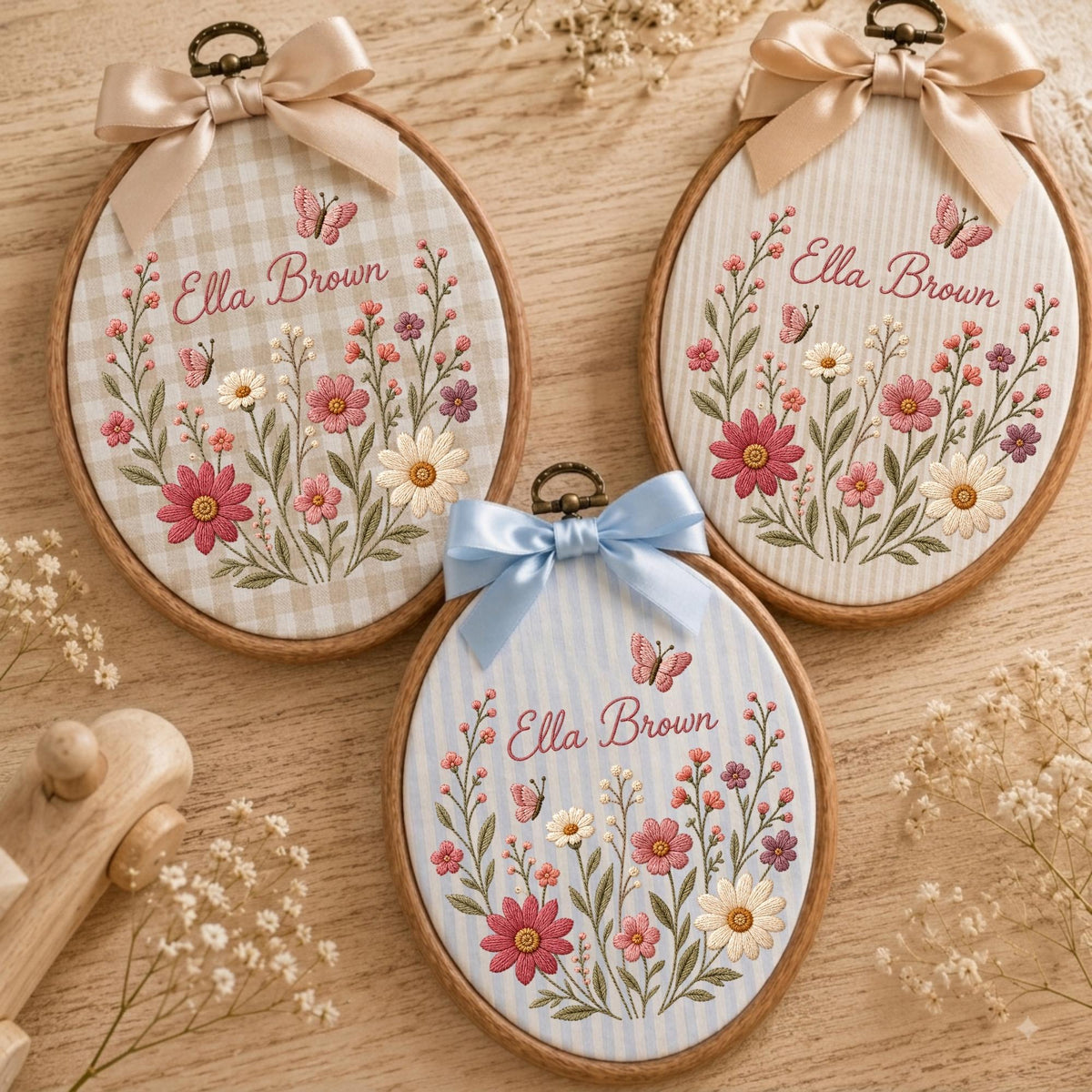 May include: Three oval wooden embroidery hoops featuring fabric backgrounds with floral and butterfly embroidery. Each hoop displays the name Ella Brown in cursive script, accented by a satin ribbon bow at the top in beige or light blue.