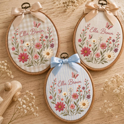 May include: Three oval wooden embroidery hoops featuring fabric backgrounds with floral and butterfly embroidery. Each hoop displays the name Ella Brown in cursive script, accented by a satin ribbon bow at the top in beige or light blue.