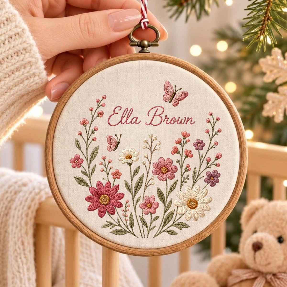 May include: A circular wooden embroidery hoop containing off-white fabric with a hand-stitched floral design. The embroidery features pink, white, and purple flowers, green stems, and two small butterflies. The name Ella Brown is stitched in pink.