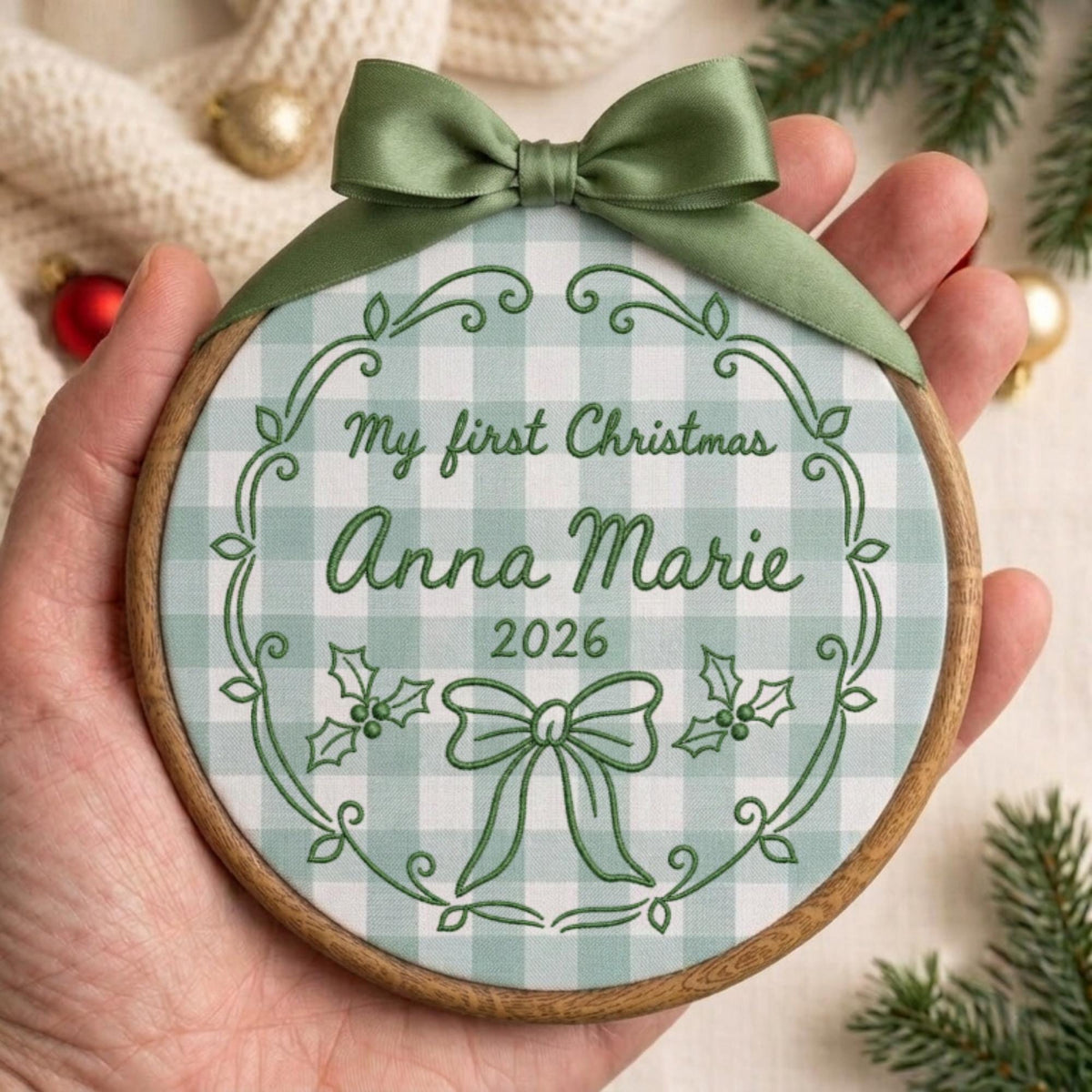 May include: A round wooden embroidery hoop ornament featuring a light green and white gingham fabric background. The fabric is embroidered with dark green text reading My first Christmas Anna Marie 2026, a decorative wreath, and a bow. A sage green satin ribbon is tied at the top.