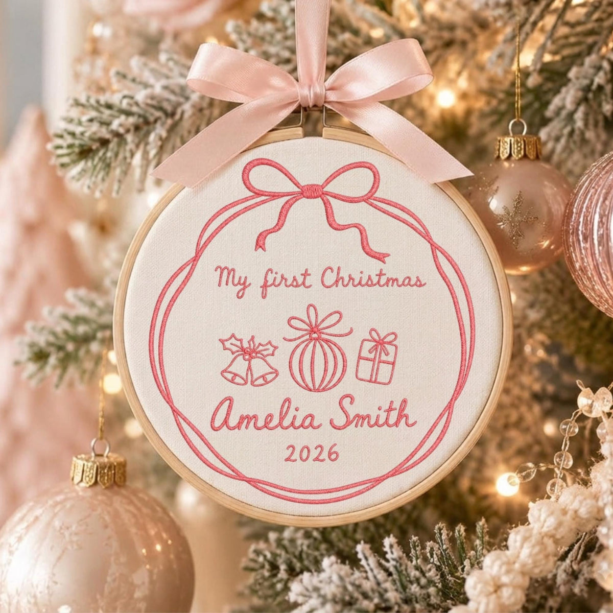 May include: A circular wooden embroidery hoop ornament featuring white fabric with pink stitched text reading My first Christmas Amelia Smith 2026, accompanied by embroidered icons of bells, a bauble, and a gift, topped with a pink satin ribbon bow.