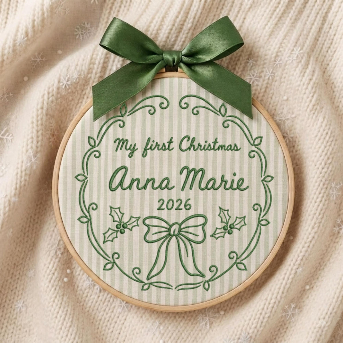 May include: A circular wooden embroidery hoop containing fabric with light green vertical stripes. The fabric features dark green embroidered text reading My first Christmas Anna Marie 2026, a decorative border, holly leaves, and a bow. A dark green satin ribbon is tied at the top.