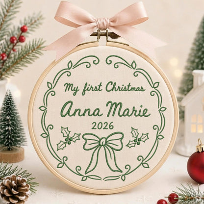 May include: A circular wooden embroidery hoop containing cream-colored fabric with green embroidered text reading My first Christmas Anna Marie 2026, surrounded by a decorative vine border, holly leaves, and a bow, topped with a pale pink ribbon.