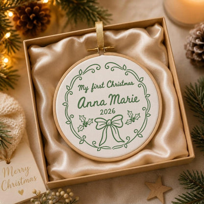 May include: A round wooden embroidery hoop containing white fabric with green embroidered text reading My first Christmas Anna Marie 2026, surrounded by a decorative green wreath, holly leaves, and a bow, presented in a gift box.