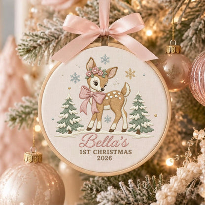 May include: A round wooden embroidery hoop ornament featuring a light brown fawn with a pink bow and flower crown, flanked by two green pine trees. The text reads Bella's 1ST CHRISTMAS 2026. A soft pink satin ribbon is tied at the top of the hoop.