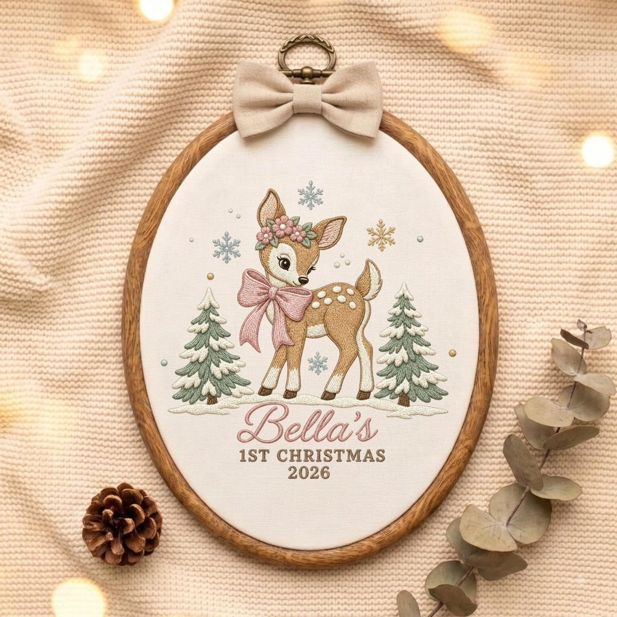 May include: An oval embroidery hoop featuring a fawn with a floral crown and pink bow, flanked by two green pine trees and snowflakes. The text reads Bella's 1ST CHRISTMAS 2026. A beige fabric bow is attached to the top of the wooden frame.