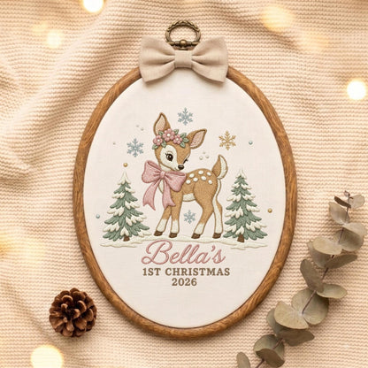 May include: An oval embroidery hoop featuring a fawn with a floral crown and pink bow, flanked by two green pine trees and snowflakes. The text reads Bella's 1ST CHRISTMAS 2026. A beige fabric bow is attached to the top of the wooden frame.