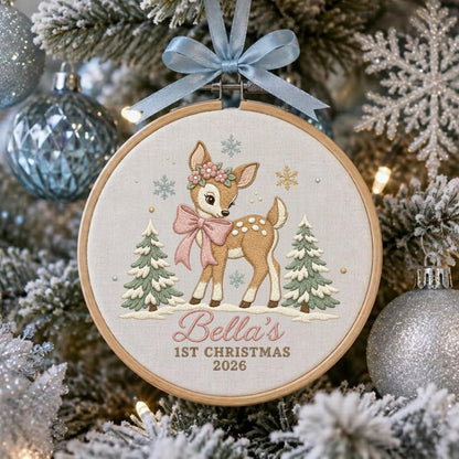 May include: A circular wooden embroidery hoop ornament featuring a light-colored fabric background with a stitched fawn wearing a pink bow and floral crown, flanked by two pine trees, with the text Bella's 1ST CHRISTMAS 2026 and a blue ribbon.