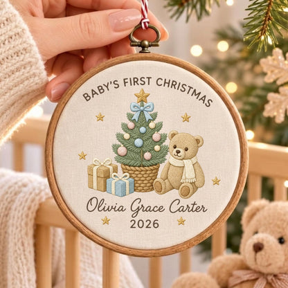 May include: A round wooden embroidery hoop ornament featuring a cream fabric center with embroidered text Baby's First Christmas, Olivia Grace Carter, and 2026. The design includes a green Christmas tree, a tan teddy bear, and gift boxes.