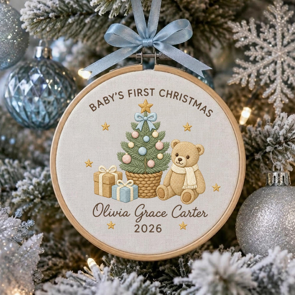 May include: A round wooden embroidery hoop ornament featuring a light-colored fabric with an embroidered Christmas tree, a teddy bear, and two gift boxes. The text reads BABY'S FIRST CHRISTMAS, Olivia Grace Carter, and 2026. A light blue ribbon bow.
