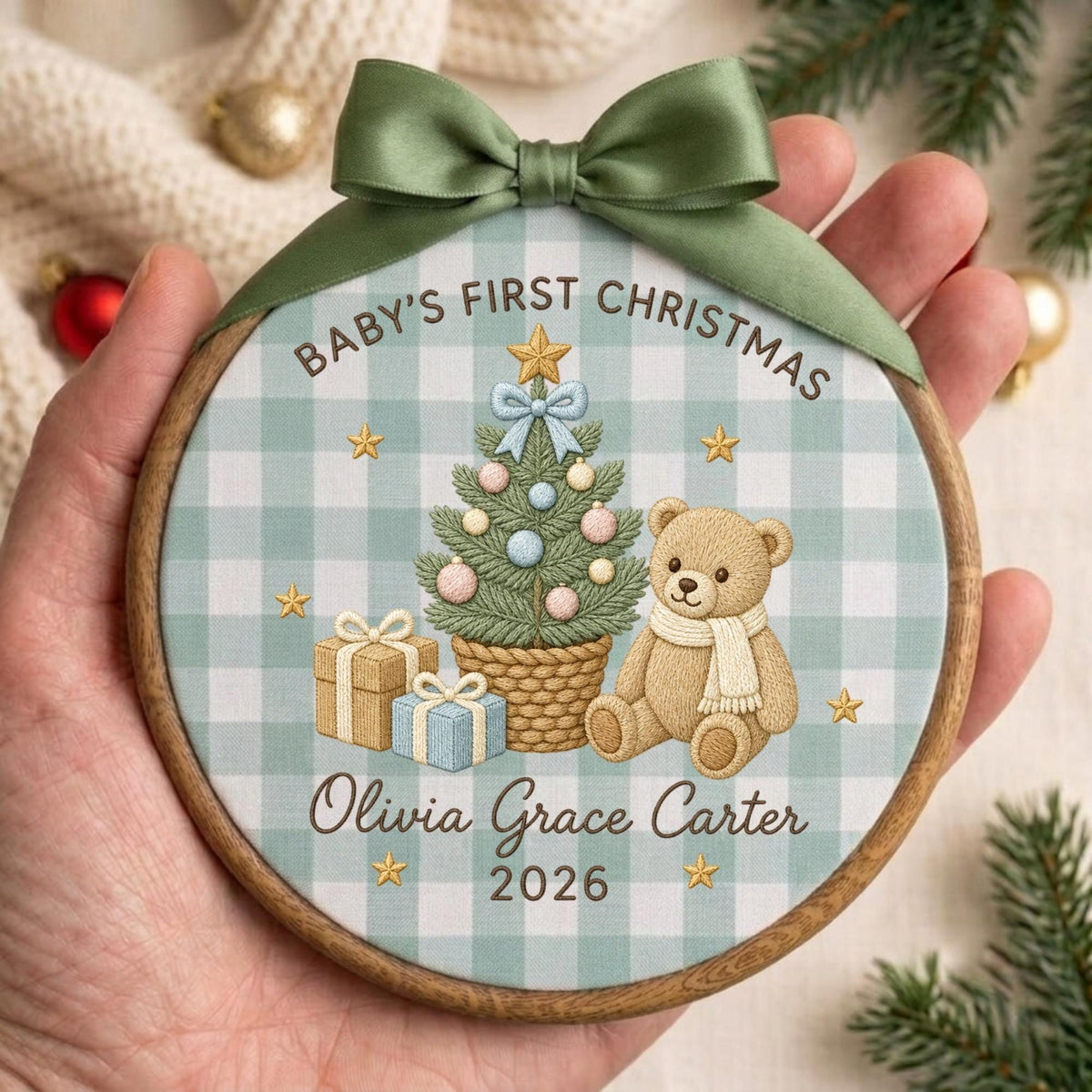 May include: A round wooden embroidery hoop ornament featuring a light blue and white gingham fabric background. The design includes a Christmas tree, a teddy bear, gifts, and gold stars. Text reads: BABY'S FIRST CHRISTMAS, Olivia Grace Carter, 2026.