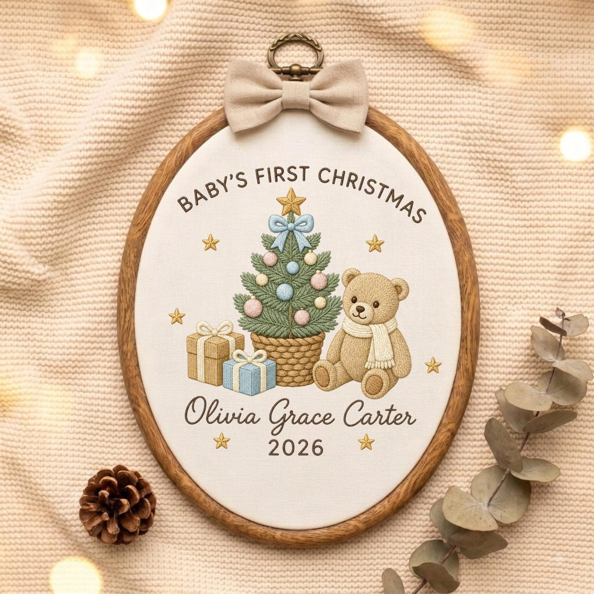 May include: An oval wooden embroidery hoop containing white fabric with embroidered text Baby's First Christmas, Olivia Grace Carter, and 2026. The design features a decorated Christmas tree, a teddy bear with a scarf, and gifts, topped with a beige bow.