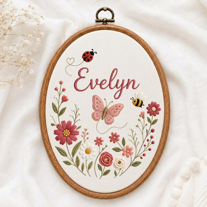 May include: An oval wooden embroidery hoop containing white fabric with the name Evelyn stitched in pink cursive. The design features a red ladybug, a pink butterfly, a yellow and black bee, and various colorful floral stems and blossoms.
