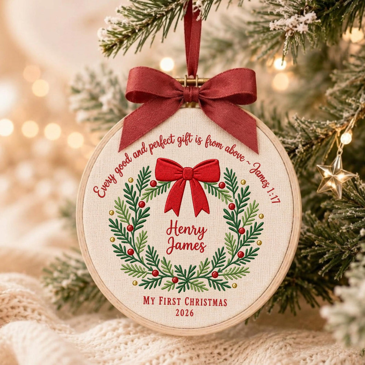 May include: A round wooden embroidery hoop ornament featuring a cream-colored fabric center with an embroidered green wreath, red berries, and a red bow. The text reads: Every good and perfect gift is from above ~ James 1:17, Henry James, and MY FIRST CHRISTMAS 2026.